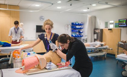 Med students practicing intubating with a dummy on a stretcher. She is bent over the head, while another woman watches